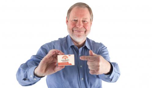 Na zdjęciu uśmiechnięty senior (jasne włosy, siwa broda, ubrany jest w niebieską koszulę), który w prawej ręce trzyma Konstancińską Kartę Seniora.