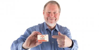 Na zdjęciu uśmiechnięty senior (jasne włosy, siwa broda, ubrany jest w niebieską koszulę), który w prawej ręce trzyma Konstancińską Kartę Seniora. 