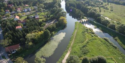 Zdjęcie miasta z lotu ptaka. W dolnej części widać ulicę i jadące po niej samochody. Obok ulicy płynie rzeka. W dali widoczne są zabudowania jednorodzinne.